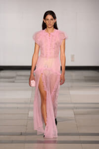 Model walking the runway in a sheer pink ruffled gown with a front slit, holding a small pink clutch.