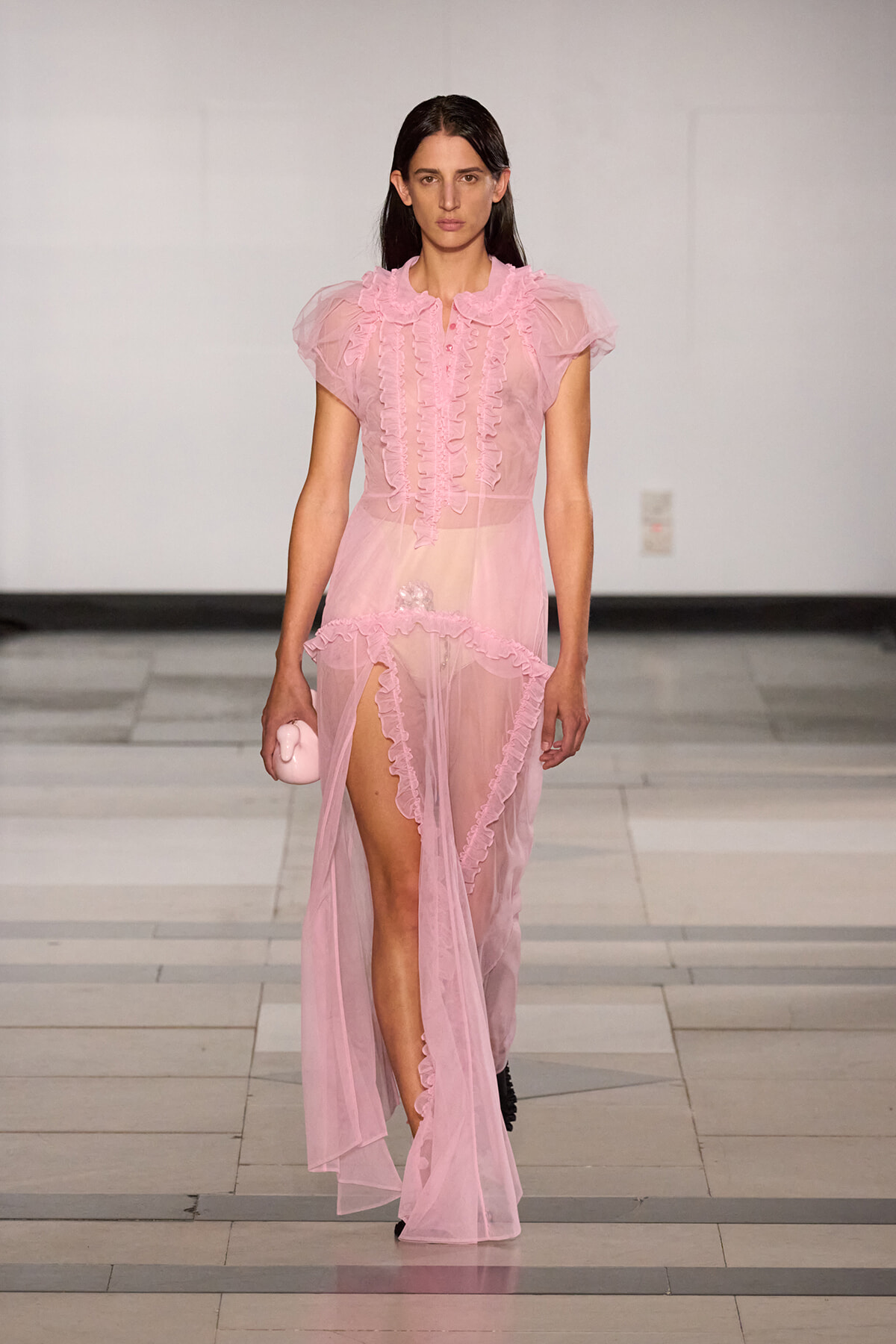Model walking the runway in a sheer pink ruffled gown with a front slit, holding a small pink clutch.