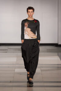 Model walking the runway in a black top with a painterly print and red flower embellishment, paired with loose black pants and flat sandals on a light-gray catwalk.