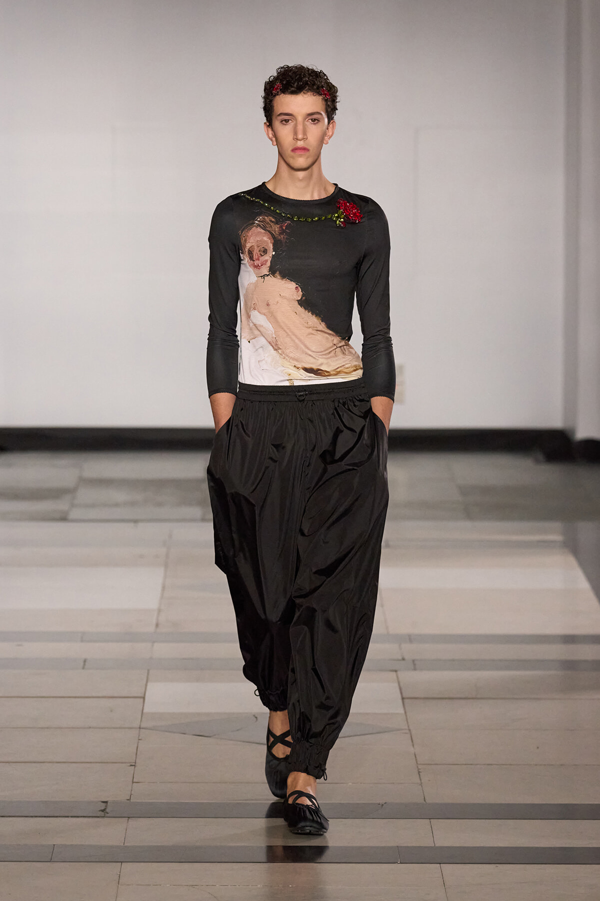 Model walking the runway in a black top with a painterly print and red flower embellishment, paired with loose black pants and flat sandals on a light-gray catwalk.