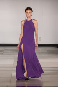 Model walking a purple halter-neck gown with a high slit on one leg, accessorized by long crystal earrings on a runway.