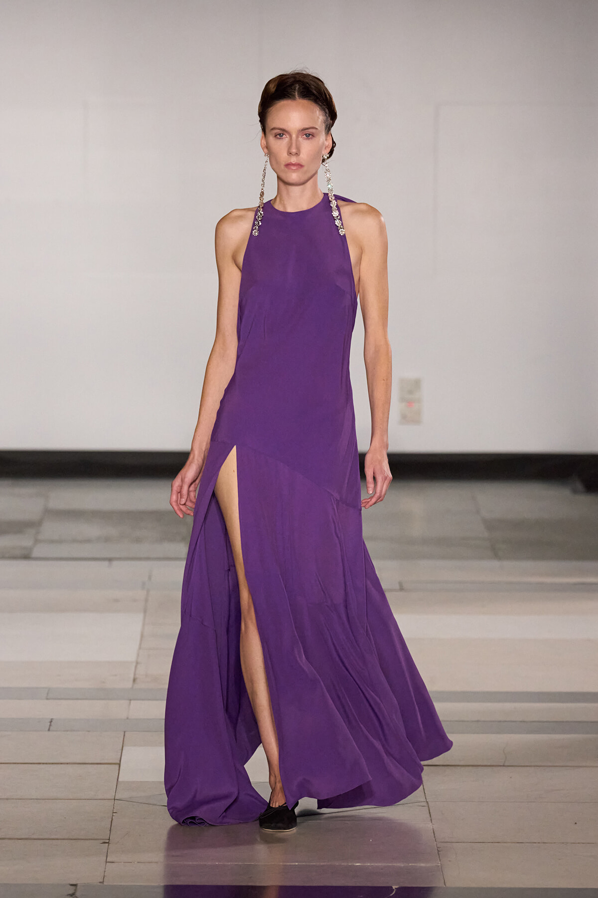 Model walking a purple halter-neck gown with a high slit on one leg, accessorized by long crystal earrings on a runway.