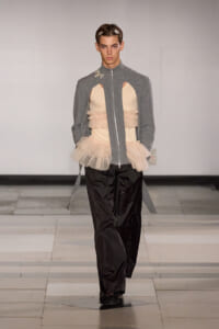 Model walking runway in a gray zip-front jacket with cutout beige corset and layered tulle peplum, paired with black wide-leg pants and small hair accessories. Concealed platform shoes.