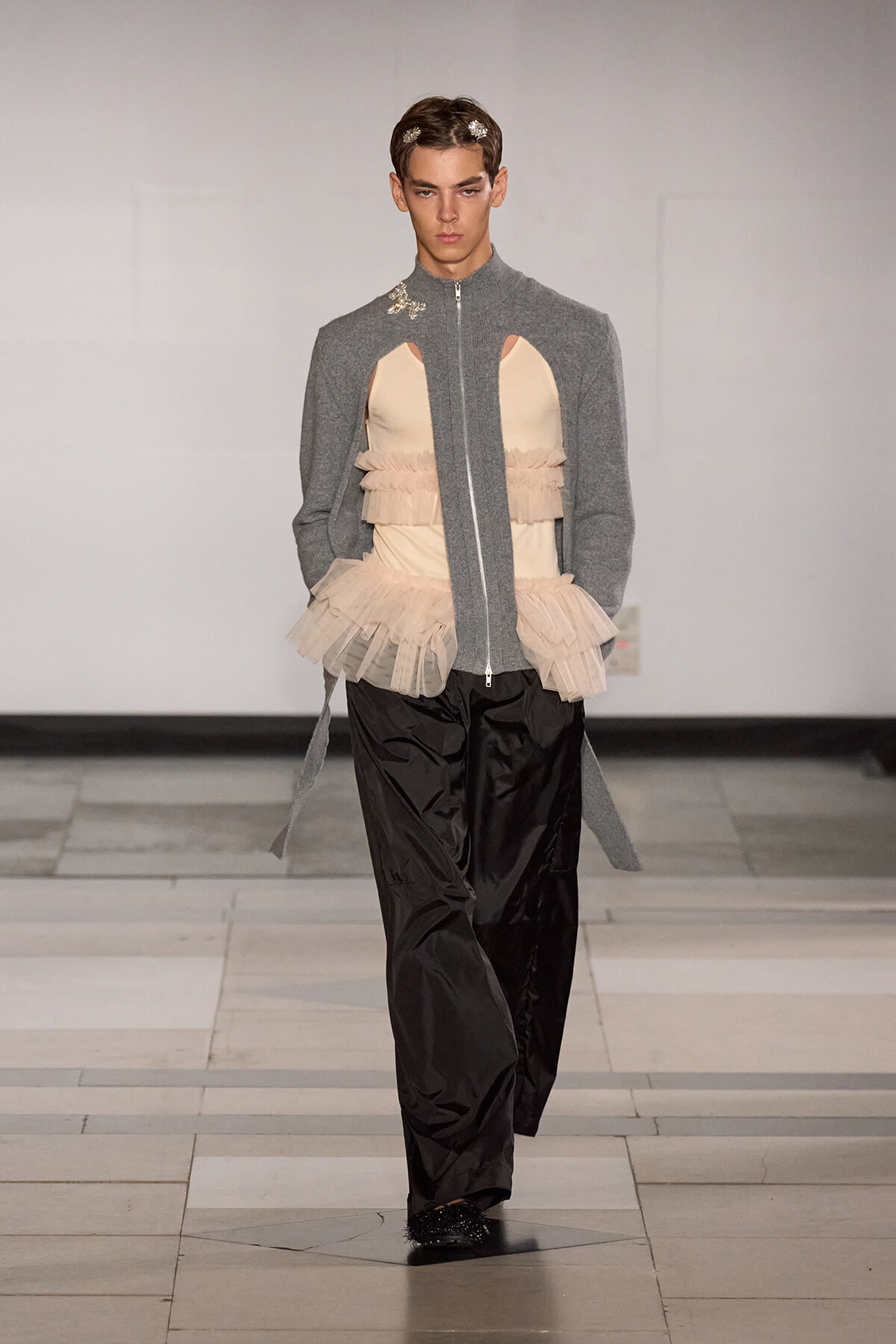 Model walking runway in a gray zip-front jacket with cutout beige corset and layered tulle peplum, paired with black wide-leg pants and small hair accessories. Concealed platform shoes.
