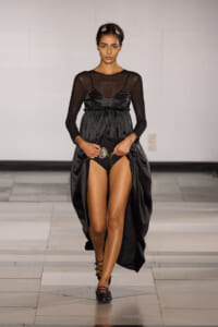 Model walking a runway in a black satin gown with sheer long sleeves and a ruched waist, stepping forward with a jeweled accessory at the waist.