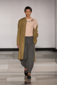 Model wearing a tan oversized trench coat over a pale pink top with a jeweled brooch, gray harem-style pants, and black sandals on a runway.