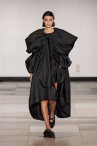 Model walking a runway in a dramatic black sculptural gown with a large bow at the neckline and voluminous sleeves, featuring a high-low draped skirt and flat, sparkly shoes.