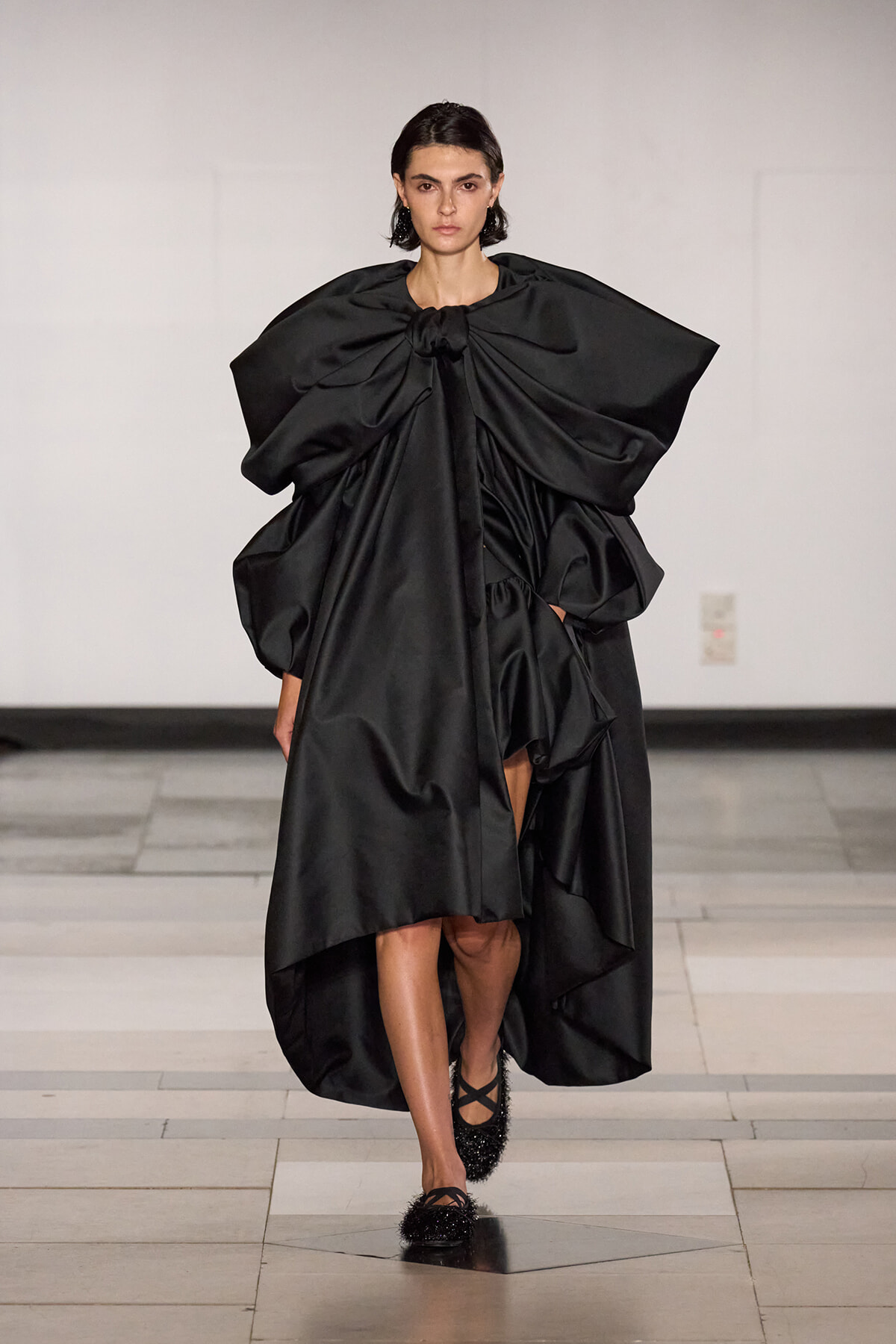 Model walking a runway in a dramatic black sculptural gown with a large bow at the neckline and voluminous sleeves, featuring a high-low draped skirt and flat, sparkly shoes.