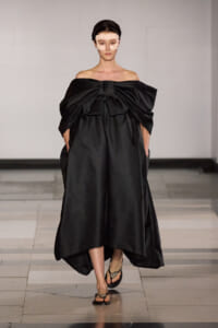 Model walks a runway in a voluminous black off-shoulder gown with a large front bow, paired with a pale fabric face mask and jeweled sandals.