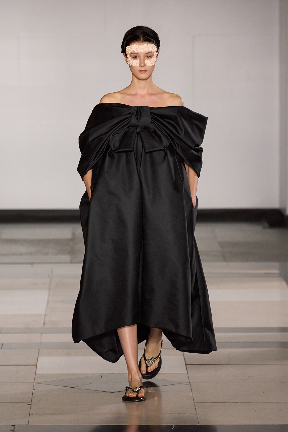 Model walks a runway in a voluminous black off-shoulder gown with a large front bow, paired with a pale fabric face mask and jeweled sandals.