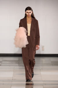 Runway model in a brown oversized suit with a beige top, left arm in pink tulle sleeve, wearing a lace mask.