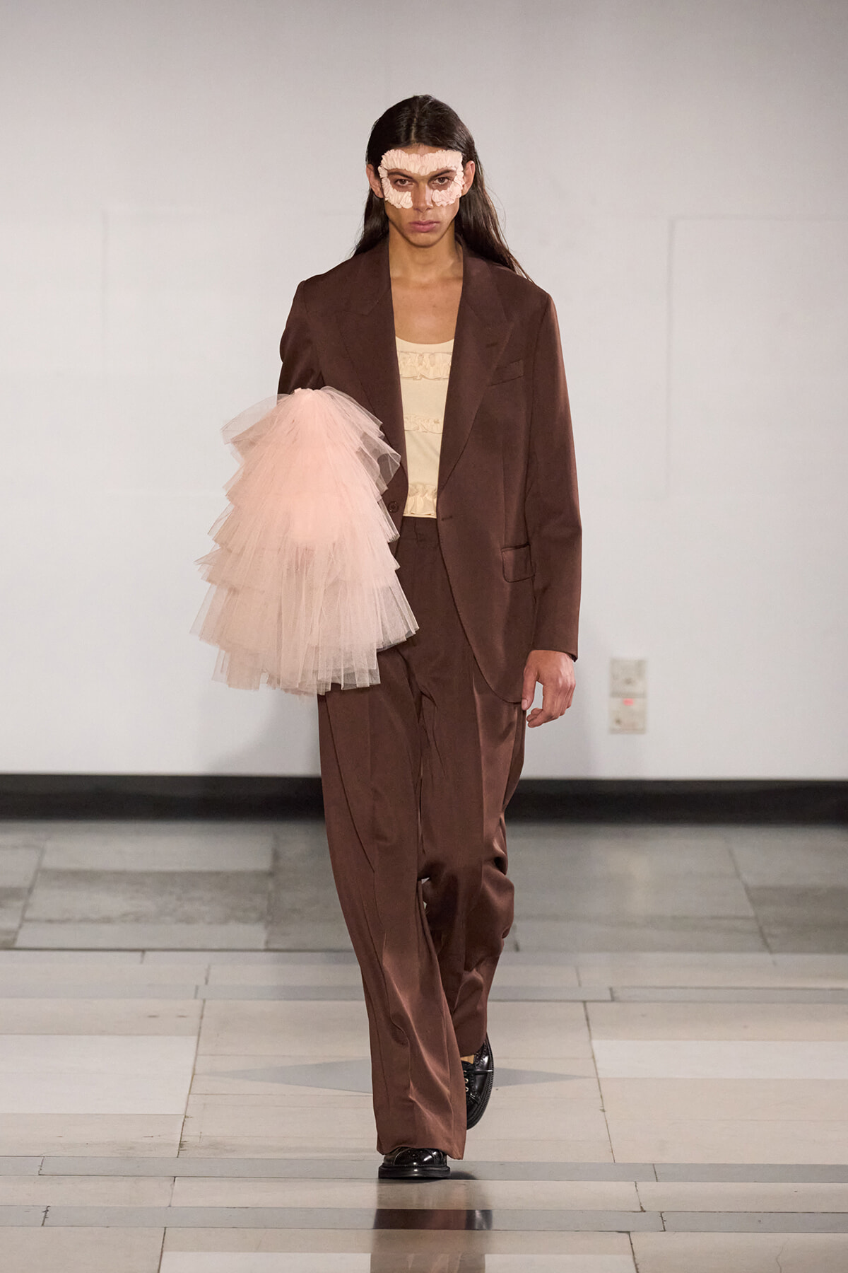 Runway model in a brown oversized suit with a beige top, left arm in pink tulle sleeve, wearing a lace mask.