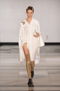 Runway model wearing an oversized white shirt dress with pearl embellishments, carrying a pearl-strand bag and a white ceramic bottle.