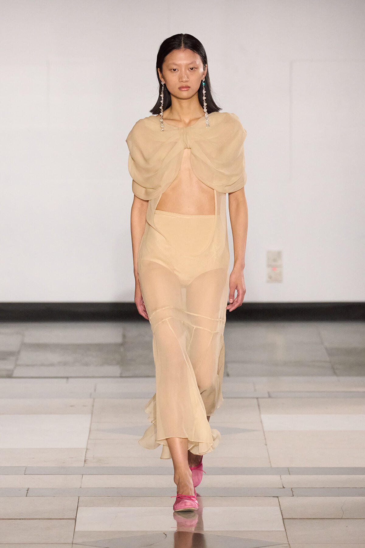 Model walks the runway in a beige sheer two-piece outfit with a draped cape-like top and pink shoes on a white backdrop runway.