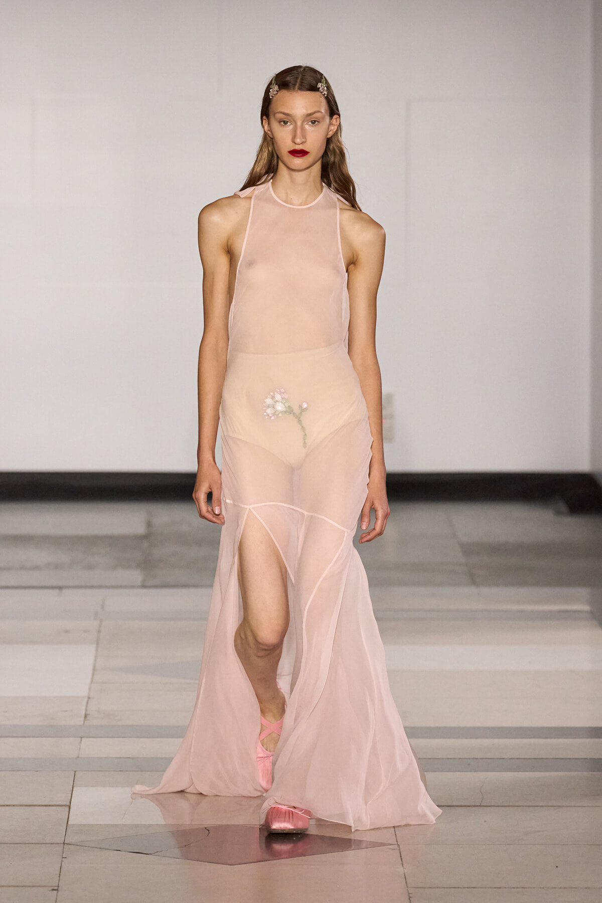 Runway model in a sheer blush-pink gown with a floral embroidery design at the hip, walking on a fashion show catwalk.