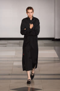 Model walking a runway in a long black coat, holding its lapel with crossed arms, wearing pink tights and black flats with a chain detail; white studio backdrop.