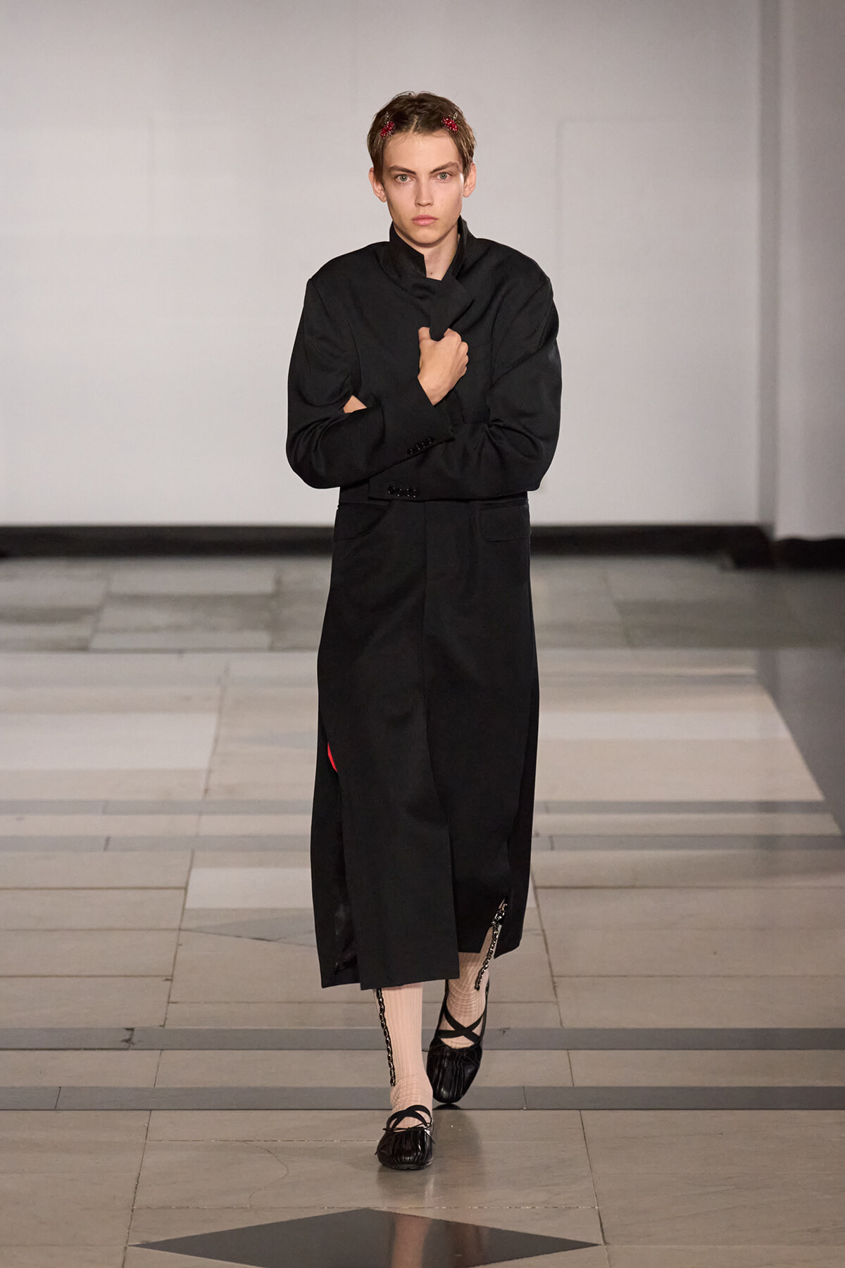 Model walking a runway in a long black coat, holding its lapel with crossed arms, wearing pink tights and black flats with a chain detail; white studio backdrop.