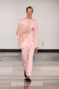 Male model in a pink suit walks the runway holding a pale pink feathered accessory. Clean, minimalist backdrop.