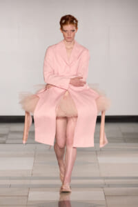 Model on a runway wearing a pale pink blazer over a pink tutu skirt with tulle peeking out, a serious expression, and hair clipped back.
