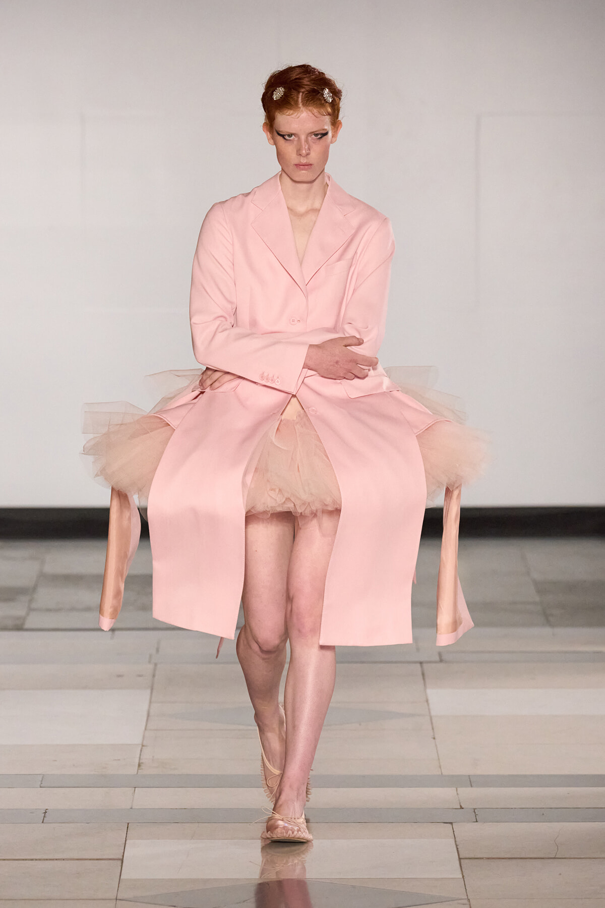 Model on a runway wearing a pale pink blazer over a pink tutu skirt with tulle peeking out, a serious expression, and hair clipped back.