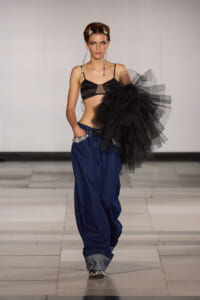 Model walks a runway in a black bralette, oversized denim trousers with jeweled pockets, and a large black tulle bow on the shoulder. Jewelry complements the look.