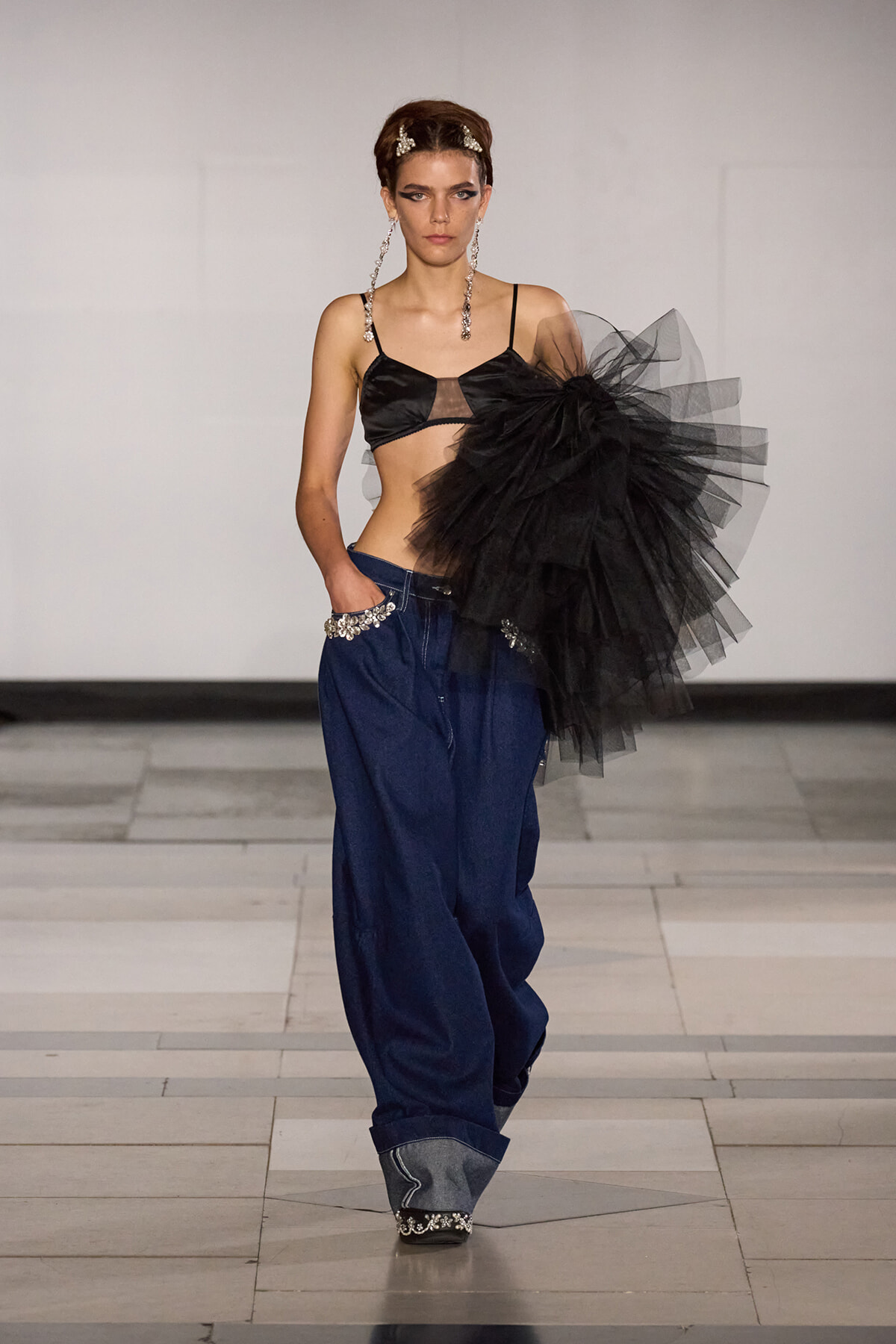 Model walks a runway in a black bralette, oversized denim trousers with jeweled pockets, and a large black tulle bow on the shoulder. Jewelry complements the look.
