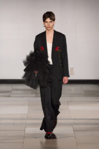 Runway look: a model in a black suit with red rose embellishments, white top, and a large black tulle piece draped over the arm.