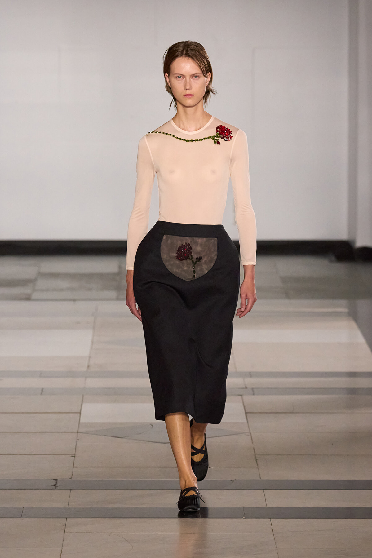 Model walks the runway wearing a sheer beige long-sleeve top with a green chain detail and red floral embellishment at the shoulder, paired with a black midi skirt featuring a decorative pocket with embroidery in front.