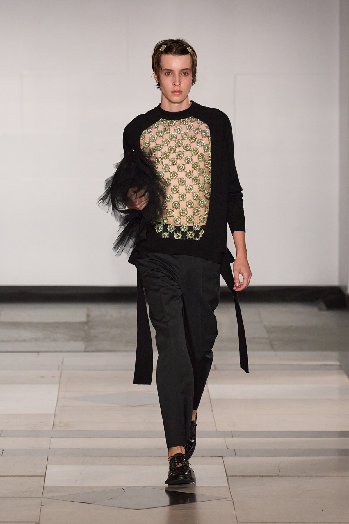 Male model on a runway wearing a black knit top with a beige jeweled mesh panel, black trousers, and a black tulle drape on the left arm, polished lace-up shoes nearby
