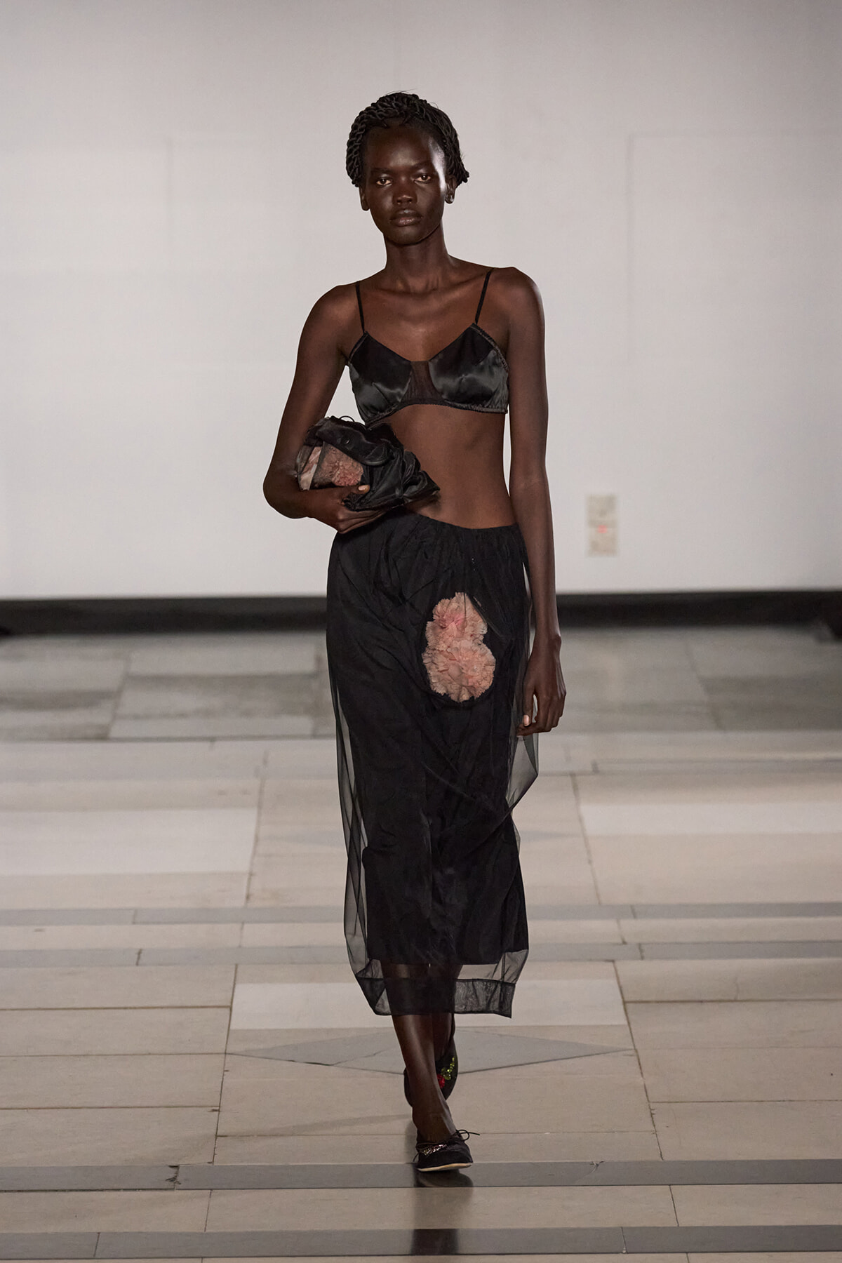 Model walks a runway in a black satin bralette and sheer black skirt with a pink floral appliqué, carrying a black clutch.