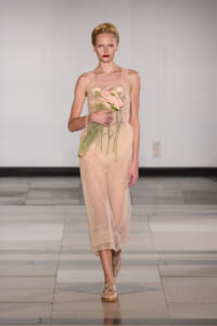 Model walking runway in a sheer nude dress, holding a bouquet of pink flowers wrapped in green tissue. Blonde hair, red lipstick, neutral backdrop.