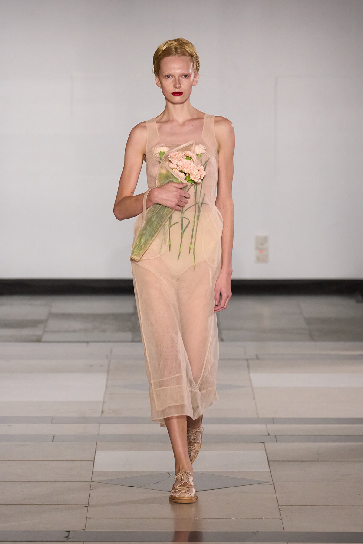 Model walking runway in a sheer nude dress, holding a bouquet of pink flowers wrapped in green tissue. Blonde hair, red lipstick, neutral backdrop.