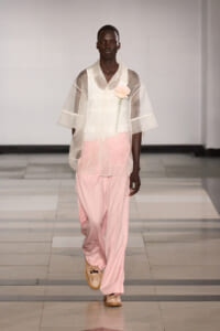 Male model walks a runway wearing a sheer white overshirt over a white shirt and pale pink wide-leg pants, with a pink flower corsage at the chest.