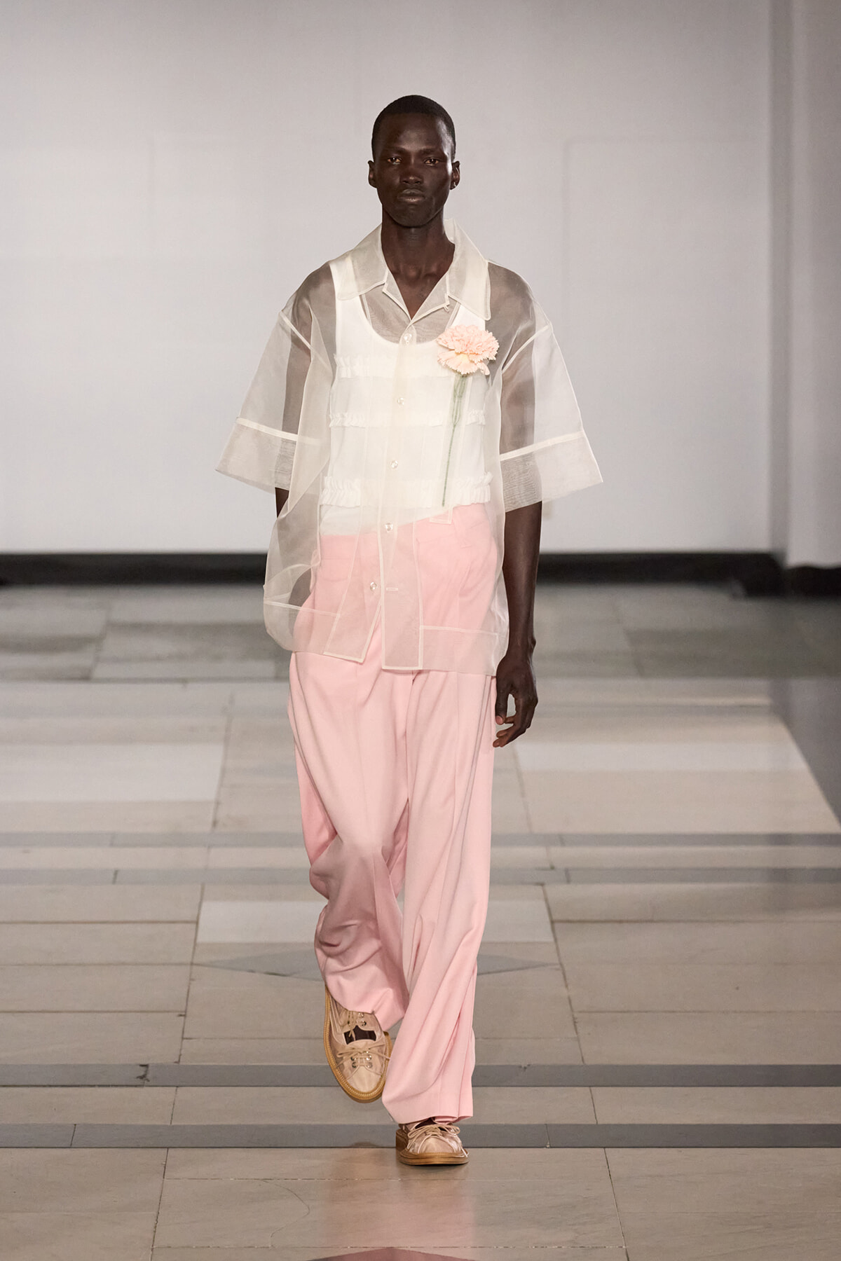 Male model walks a runway wearing a sheer white overshirt over a white shirt and pale pink wide-leg pants, with a pink flower corsage at the chest.