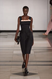 Black knee‑length dress with pink ruffle neckline on a model walking a runway, holding a small black clutch.