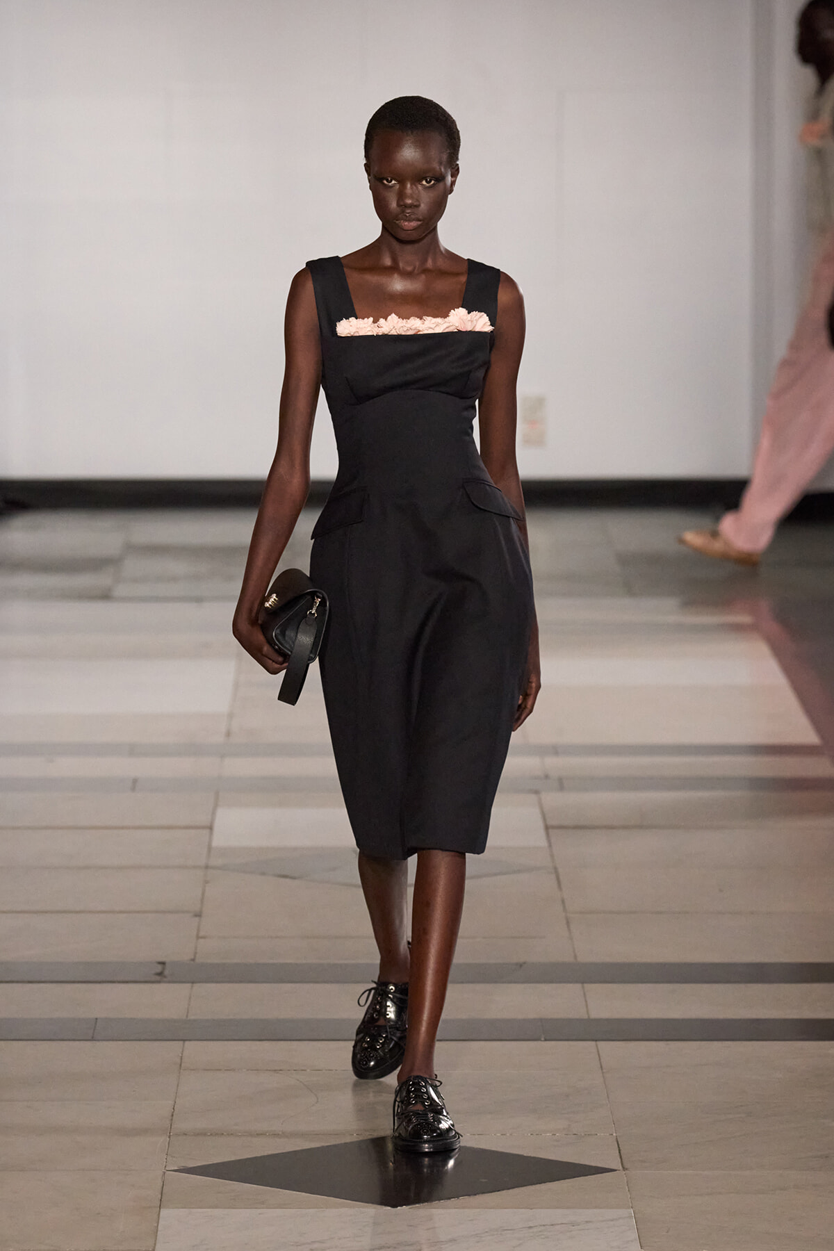 Black knee‑length dress with pink ruffle neckline on a model walking a runway, holding a small black clutch.