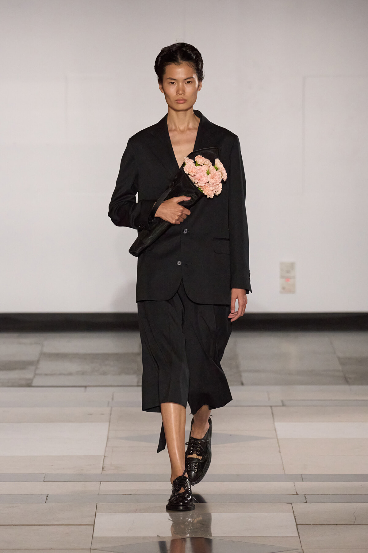 Model wearing a black blazer and wide-legged skirt on a runway, holding a bouquet of pink carnations.