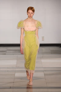 Model walks a runway in a lime-green dress with a pink lace bodice and sheer tulle sleeves on each shoulder.
