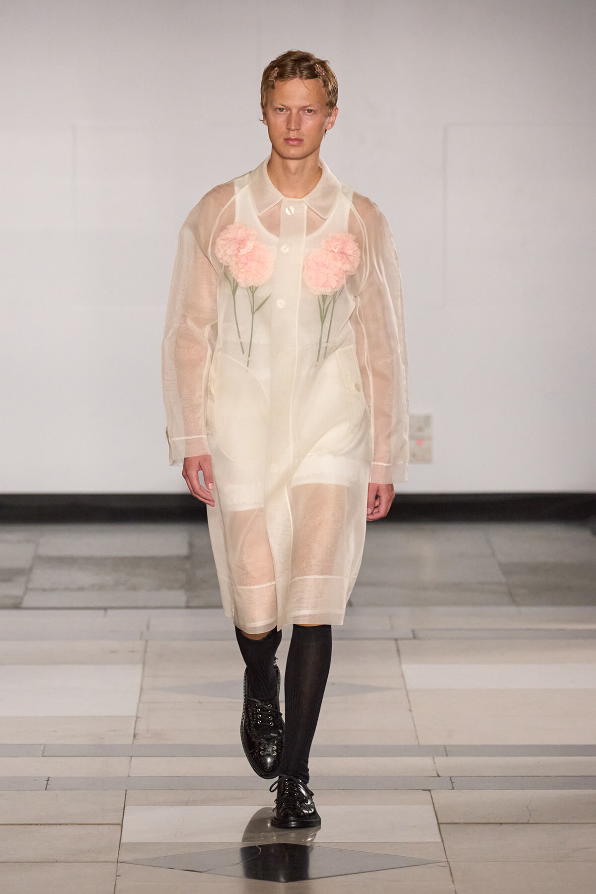 Model on a runway wearing a sheer ivory coat with pink flower appliqués on the chest, paired with black tights and shiny black shoes.