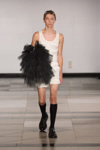Male model on a runway wearing a white sleeveless ruffled romper, black knee-high socks, and black patent shoes, carrying a large black tulle accessory.