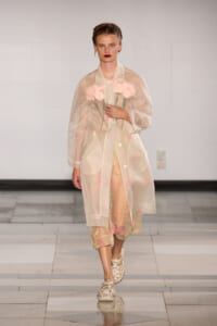 Model walks a fashion runway wearing a sheer pink-tinted coat with pink carnations tucked inside, neutral backdrop behind.