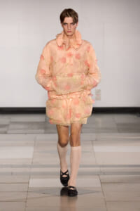 Male model walks a fashion show in a peach sheer floral jacket and matching shorts, with cream knee-high socks and black ballet flats
