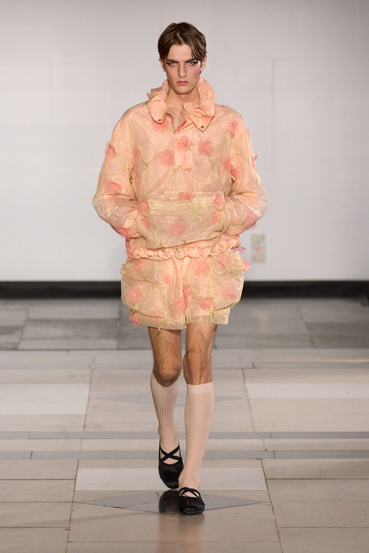 Male model walks a fashion show in a peach sheer floral jacket and matching shorts, with cream knee-high socks and black ballet flats