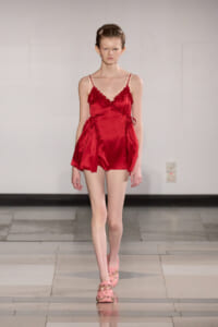 Model walks the runway in a red satin romper with ruffled trim and spaghetti straps.