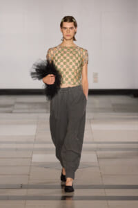 Model on a runway wearing a sheer beaded top with green floral embellishments and loose gray pants, holding a black tulle accessory.