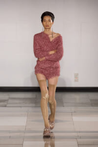 Model walking the runway in a pink fuzzy knit romper with an off-shoulder neckline, paired with beige patterned thigh-high stockings and embellished sandals.