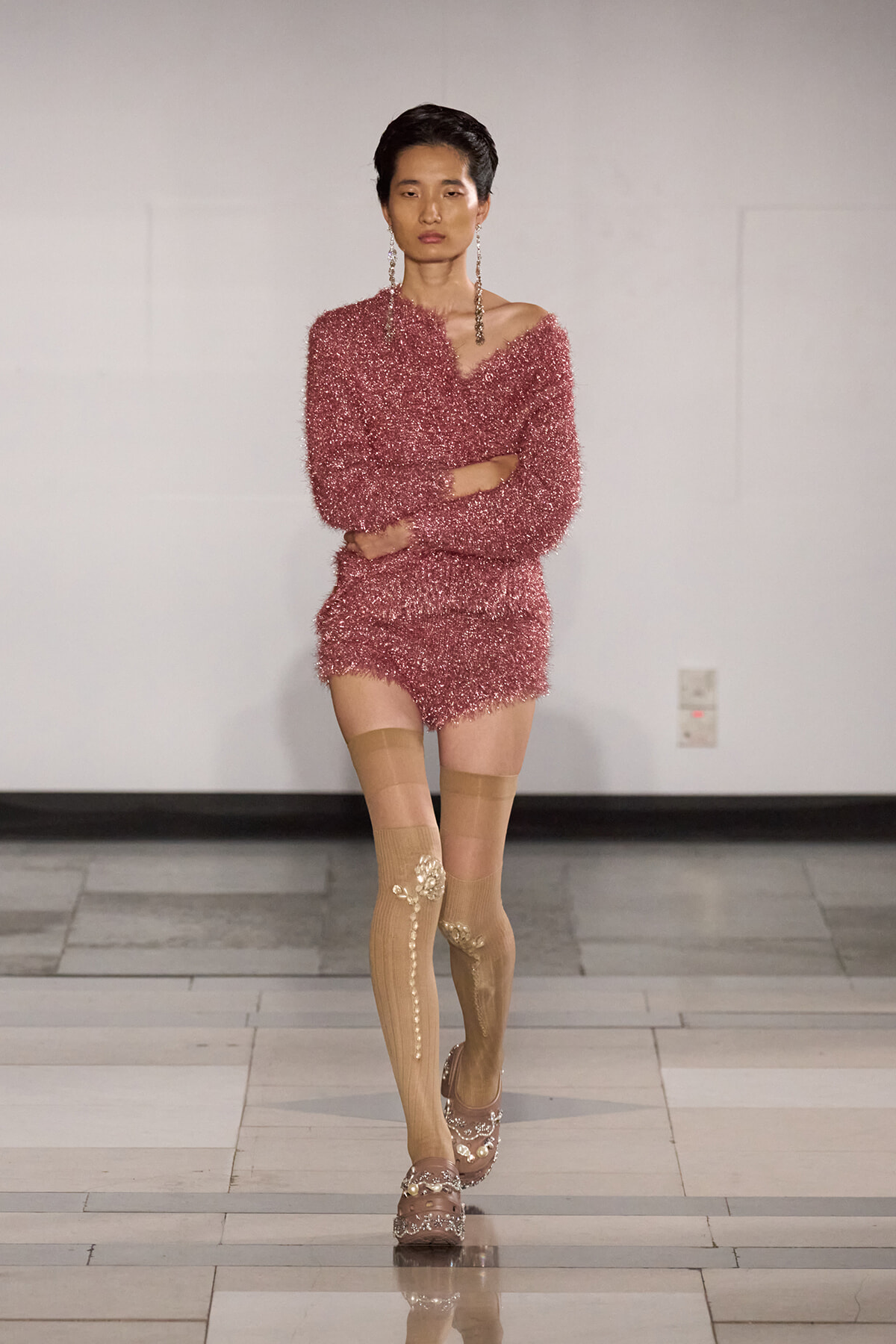 Model walking the runway in a pink fuzzy knit romper with an off-shoulder neckline, paired with beige patterned thigh-high stockings and embellished sandals.