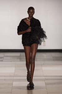 Model on a runway wearing a black sequined mini dress with a voluminous black tulle detail on the right arm, necklace included.