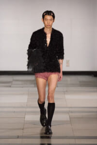 Model on a runway wearing a black glittery textured jacket with matching shorts, black knee-high socks, and shiny black lace-up shoes, holding a black tulle accessory on his arm.