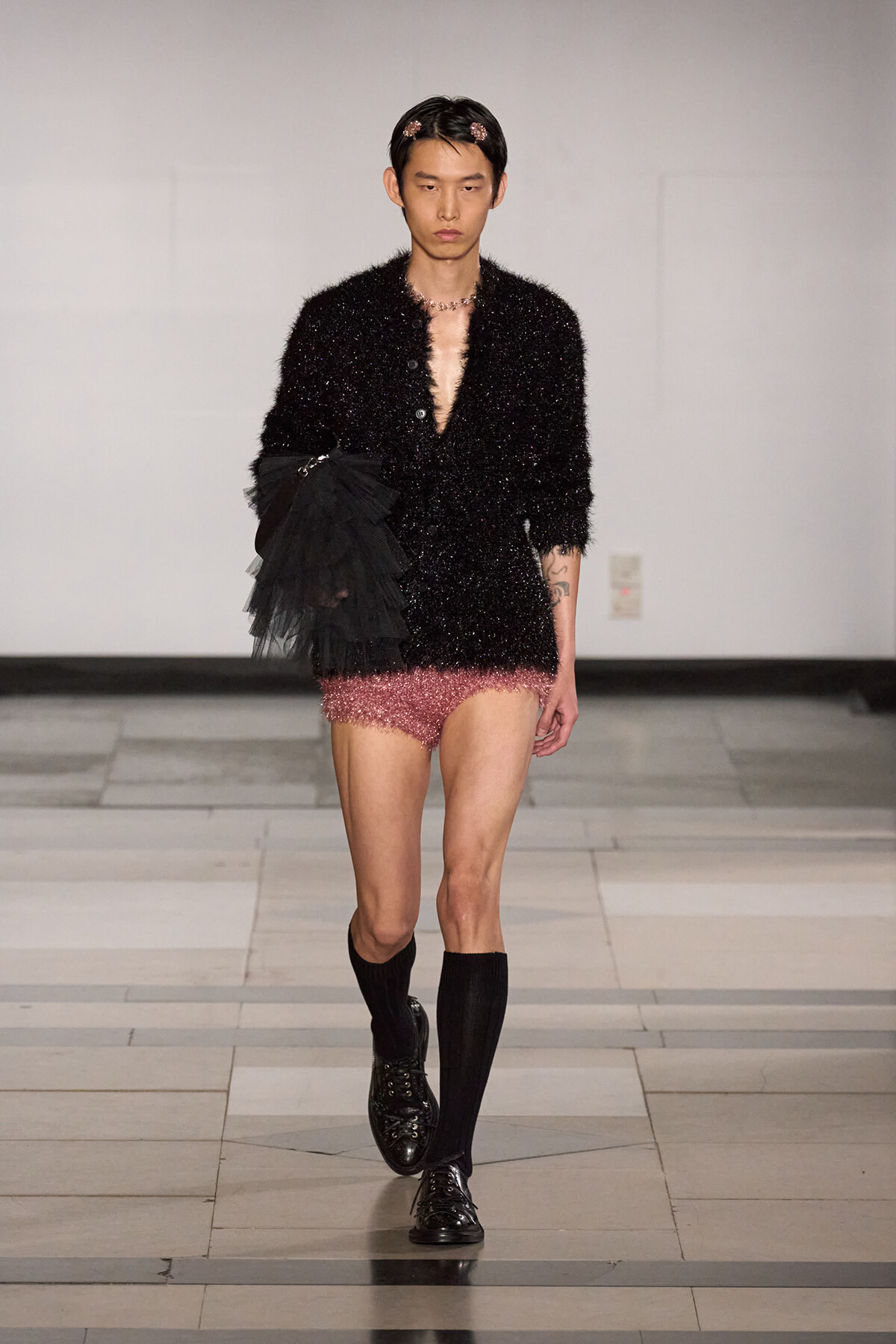 Model on a runway wearing a black glittery textured jacket with matching shorts, black knee-high socks, and shiny black lace-up shoes, holding a black tulle accessory on his arm.